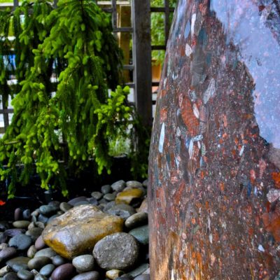 Large polished stone water feature on smooth river rocks, with green foliage and a wooden trellis in the background.