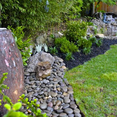 Landscaped backyard with a stone water feature, smooth river rocks, green plants, and a small waterfall in the background.