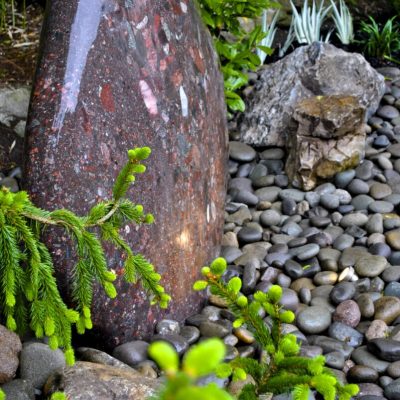Large stone water feature surrounded by smooth river rocks, green foliage, and a few large stones in a garden setting.