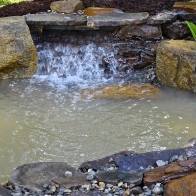 A small backyard pond with a stone waterfall feature and green plants at the water’s edge.