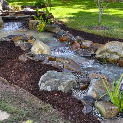 A small artificial stream with rocks and plants flows through a landscaped garden with mulch and grass.