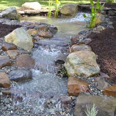 A small artificial stream with rocks and plants flows into a pond in a landscaped garden.