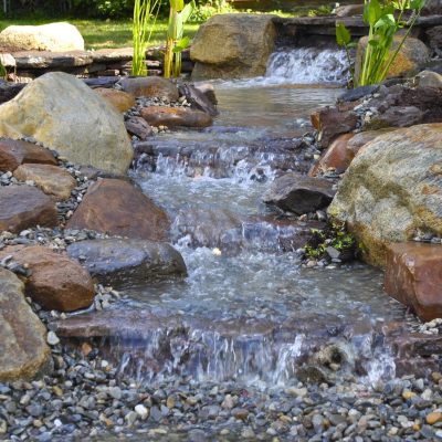 A small artificial stream flows over rocks and pebbles in a landscaped garden with surrounding plants and mulch.