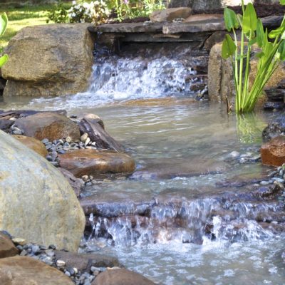 Small waterfall cascading over rocks in a landscaped garden pond with green plants and pebbles around the water.