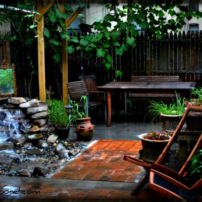 A backyard patio with wooden furniture, a small waterfall feature, potted plants, and a wooden privacy fence.