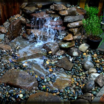 Small backyard waterfall and pond with rocks, potted plants, and a decorative clay pot next to a wooden fence.