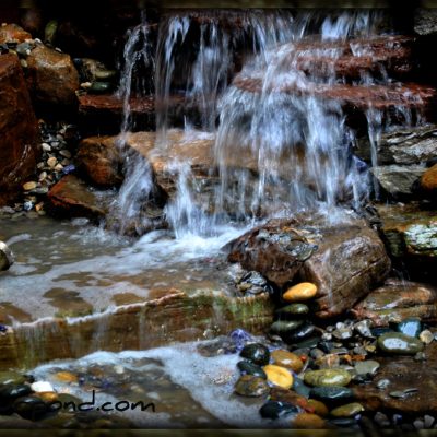 Small waterfall flowing over rocks into a shallow pool with multicolored stones and a metal cup on the left.
