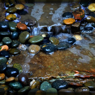 Small waterfall flowing over rocks and pebbles, surrounded by smooth, multicolored stones.