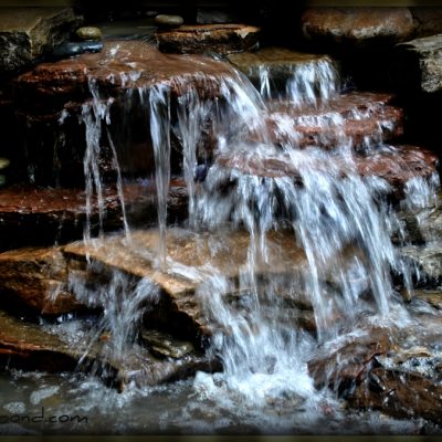 Water cascades over layered rocks in a small garden waterfall, surrounded by stones and pebbles.