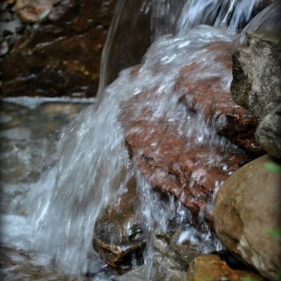 Water flows over rocks in a small waterfall, creating splashes and movement among brown and gray stones.