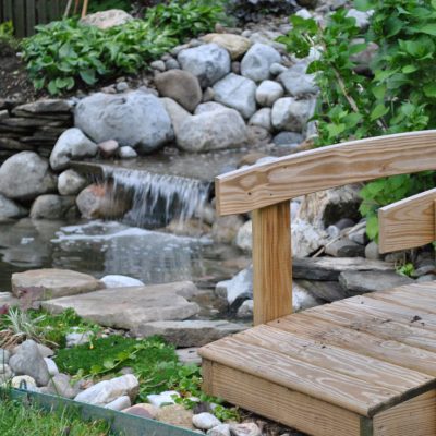 Small wooden footbridge beside a garden pond with a stone waterfall and surrounding greenery.