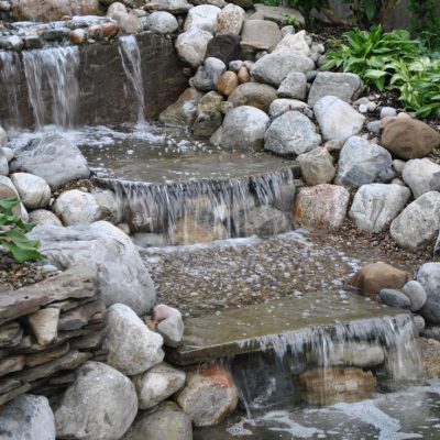 Water cascades down tiered stone steps surrounded by rocks and green plants in a landscaped garden.