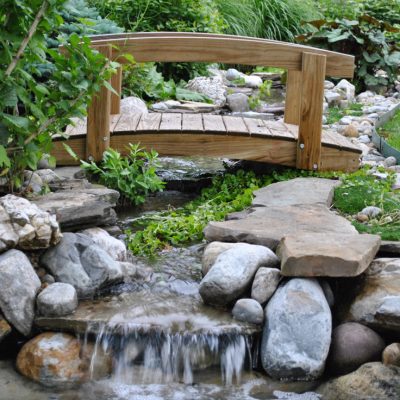 A small wooden footbridge crosses a stream with rocks and plants in a landscaped garden.
