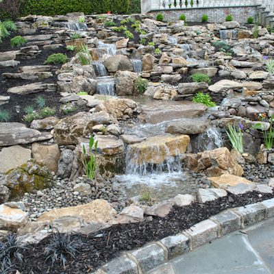 Stone waterfall feature with cascading water, surrounded by rocks, plants, and landscaped garden beds on a slope.