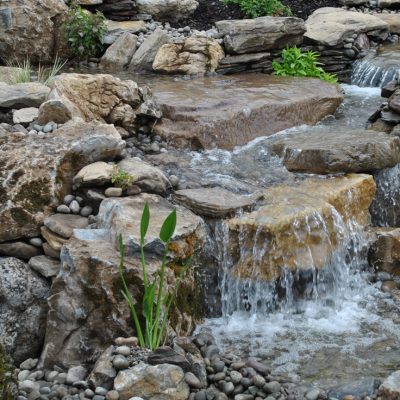 Small waterfall flowing over rocks surrounded by plants and pebbles in a landscaped garden setting.
