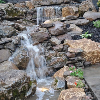 Small waterfall cascades over rocks in a landscaped garden with greenery and stone path nearby.