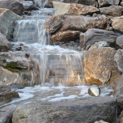 Small waterfall flowing over rocks with a metal cup partially submerged at the bottom right of the image.
