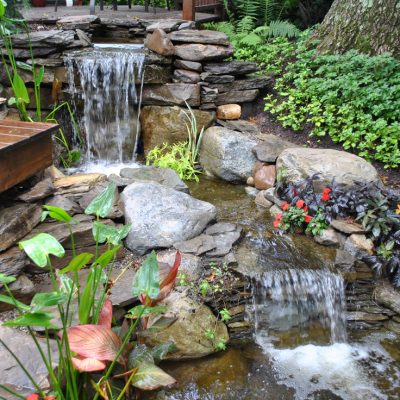 Small backyard pond with two stone waterfalls, wooden steps, surrounding plants, and blooming flowers.