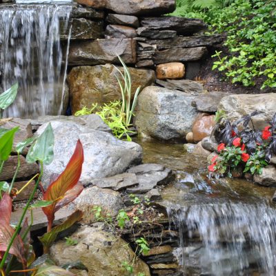 Small twin waterfalls flow over rocks into a garden pond, surrounded by green plants and red flowers.