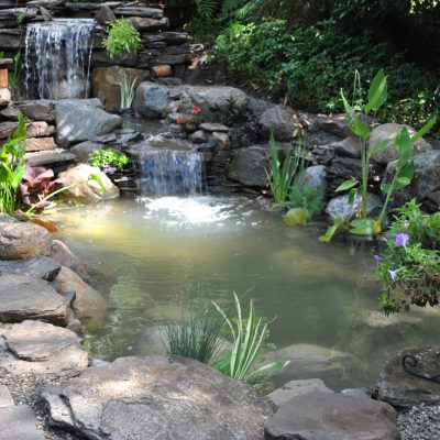 A small pond with rocks, plants, and two waterfalls beside a stone path in a garden setting.