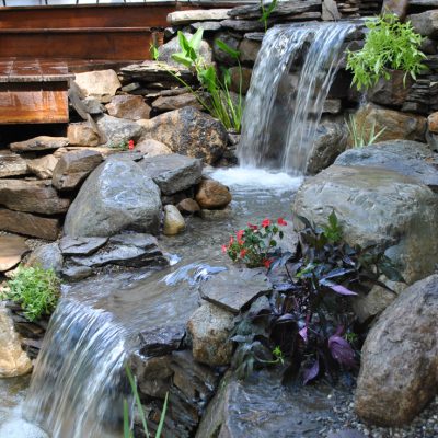 Small artificial waterfall flows over rocks in a landscaped garden surrounded by plants and flowers.