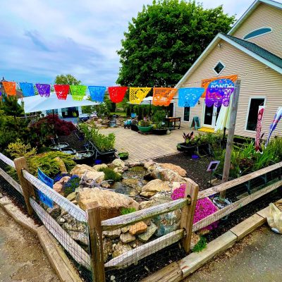 A fenced garden area with rocks, a pond, and colorful papel picado banners outside a tan building on a cloudy day.