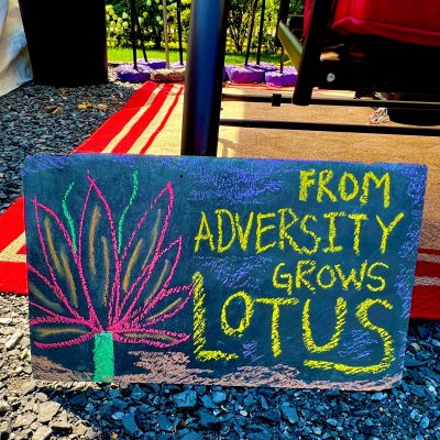 A chalkboard sign reads "From adversity grows lotus," next to a drawing of a lotus flower on gravel by a red mat.