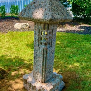 A stone lantern with a pagoda-style top stands on a grassy lawn near a greenhouse and trees.
