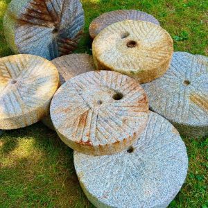Several round, textured millstones are stacked on grassy ground in bright sunlight.