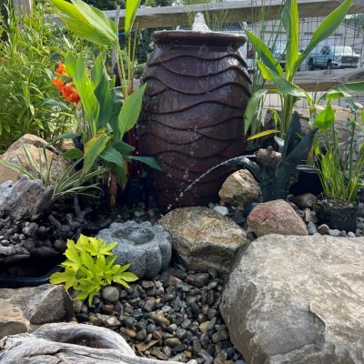 A ceramic fountain surrounded by plants, rocks, and driftwood in a landscaped garden.