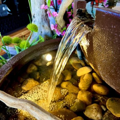 Water flows from a brown ceramic pot into a stone-filled basin, with branches and pink flowers nearby.
