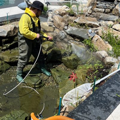 A person in waterproof gear uses a pressure washer to clean rocks in a landscaped outdoor pond.