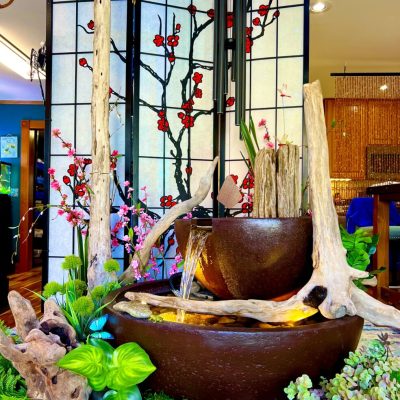 Decorative indoor water fountain with brown bowls, driftwood, green plants, and a floral screen in the background.