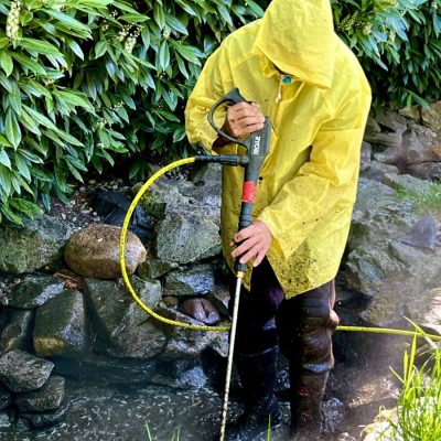 Person in a yellow raincoat pressure washes rocks near a hedge and grass in an outdoor garden area.
