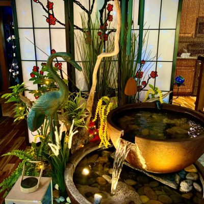 Indoor water feature with a bronze crane, ceramic lantern, plants, and a decorative stained glass backdrop with floral designs.