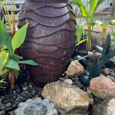A large, textured ceramic urn water feature is surrounded by rocks, plants, and a small stone basin.