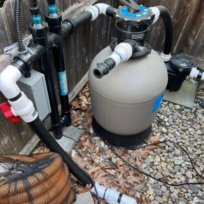 A pool filter system with pipes, valves, and a pump is installed on a bed of rocks next to a wooden fence.