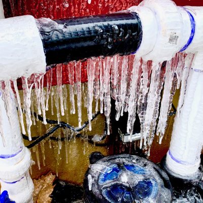 Icicles hang from frozen water pipes and valves against a red wall, indicating freezing weather conditions.
