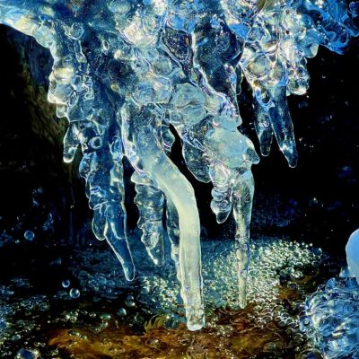 Close-up of clear, jagged icicles hanging over a shallow stream with pebbles visible beneath the water.
