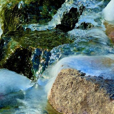 Close-up of water flowing over rocks, partially covered with ice, in bright natural light.