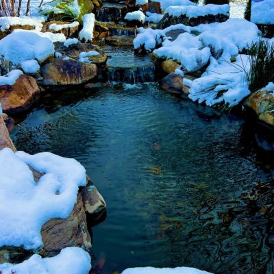 Small waterfall flows into a pond surrounded by rocks, all lightly covered with snow in a landscaped garden.