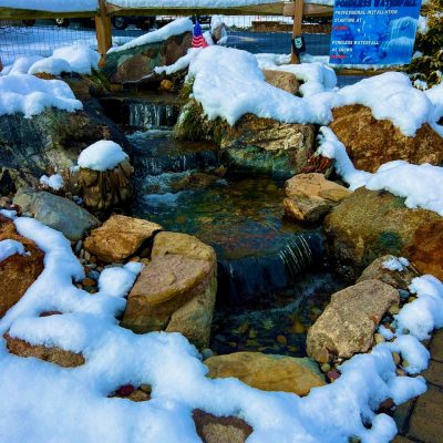 Small waterfall and rocks partially covered in snow with a sign and fence in the background.