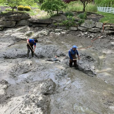Two people stand in a muddy, drained pond using shovels to clear debris, surrounded by rocks and greenery.