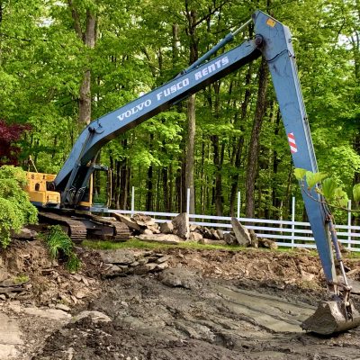 A blue excavator is digging earth near a rocky area with trees and a white fence in the background.