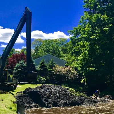 An excavator moves dirt near a riverbank while two workers stand by the water, surrounded by trees and a house.