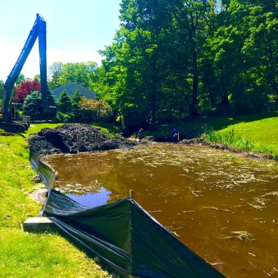 Excavator dredging a pond near trees and grass, with a black barrier along the edge and workers standing by the water.