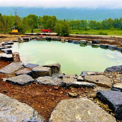 A large, circular stone pond under construction, surrounded by machinery and trees in a rural outdoor setting.