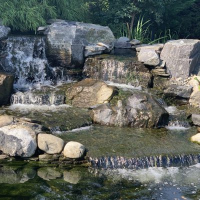 Small waterfall flows over rocks into a pond, surrounded by stones and green plants in a garden setting.