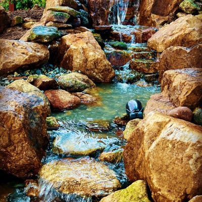 A small artificial waterfall flows over rocks into a pond, surrounded by stones and greenery.
