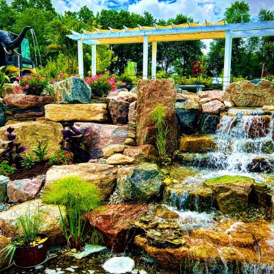 Multi-level rock waterfall in a landscaped garden with flowers, plants, and a black elephant statue in the background.